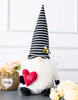 Load image into Gallery viewer, Lifestyle picture of the Valentine bee gnome. The gnome stands on a gray-colored table, and the wall behind it is white. Behind the gnome, on the left side, there&#39;s a basket with light peach-colored flowers. In front of the gnome, on the right side, there are cream-colored flowers resting on the table&#39;s surface.
