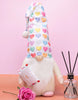 Load image into Gallery viewer, Lifestyle picture of the heart-themed gnome. Placed at a frontal angle, it stands on a peach table with a pink-colored wall behind. On the right side, a glass reed diffuser with black sticks is placed. Lastly, in front of the gnome, there&#39;s a pink flower resting on the table.
