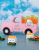 Load image into Gallery viewer, Lifestyle picture of the pink decorative Easter truck. It is positioned at a frontal angle, slightly tilted to the right, and placed on a blue table in front of a wall with a sky-themed wallpaper.
