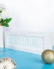 Load image into Gallery viewer, Lifestyle picture of the wooden spring sign. It is positioned at a frontal angle, but slightly tilted to the right. It is standing on a cyan-colored table, placed against a white wall. Next to the item, on the left, there&#39;s a large white flower vase with white flowers arranged in it. In front of the plaque, but to the left, a glittered gold-colored ornament can be appreciated. Lastly, positioned in the foreground, there&#39;s a light gold-colored ornament.

