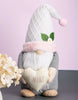 Load image into Gallery viewer, Lifestyle photo of the gnome. It is positioned at a frontal angle, standing on a gray table. The wall behind is pink. In front of the gnome, to the left, an arrangement of beige-colored flowers can be appreciated.
