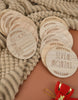 Load image into Gallery viewer, Lifestyle picture of baby milestone plaques. It presents two arrays of plaques on a cream-colored textured tablecloth.
