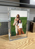 Load image into Gallery viewer, Lifestyle picture of the clear picture frame, captured from a slight left angle and standing on an unfinished wooden tabletop. A gray wall can be seen in the background. The frame holds a photo of a family: a man with his daughter on his shoulders and his wife in front of him. The picture serves as an example of the types of photos that can be placed inside.
