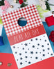 Load image into Gallery viewer, Lifestyle picture of the American picnic-themed board. It is placed at a left-tilted angle, resting on a surface divided into white, red, and blue sections. To its left, three  blue bean bags are visible, while three  red bean bags can be seen to its right.
