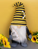 Load image into Gallery viewer, Lifestyle picture of the bee-themed gnome. It is positioned at a frontal angle, standing on a gray table. A yellow wall is positioned on the background. Behind the gnome, to the left, a bouquet of sunflowers rests on the table. In front of the gnome, to its right, another bouquet of sunflowers can be seen in a blurred view.
