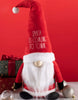 Load image into Gallery viewer, Lifestyle picture of the Christmas Santa gnome. It is placed between two gift boxes in its right and two Christmas ornaments on its left. It has a red wall behind it, and it is placed on a white table.
