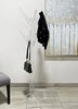 Load image into Gallery viewer, A lifestyle picture of the coat rack shows it standing on a warm gray tiled floor, positioned near a white wall. A vintage-style wall painting of a white fence covers the upper right quadrant of the wall. Next to the rack, a gray chair is partially visible, with half of its seat and one wooden leg showing. The rack holds a black coat on the upper right hook and a small black purse on the left hook below.
