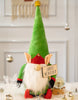 Load image into Gallery viewer, Lifestyle picture of the plush holiday gnome. Positioned at a frontal angle and standing on a table covered with a white faux fur tablecloth, the gnome is surrounded by a few champagne gold-colored Christmas ornaments. Behind the gnome, to its right, a snow globe with a wooden base can be seen. In a blurred background, a white door is visible.
