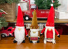 Load image into Gallery viewer, Lifestyle picture of the family of gnomes. They are placed on a wooden table in this order: Mr. Claus on the right, reindeer-themed gnome on the center, and Mrs. Claus on the left. Behind them, there&#39;s a red decorative truck carrying a Christmas tree.
