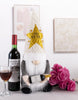 Load image into Gallery viewer, A lifestyle picture of the festive gnome is shown. The gnome is placed on a table, with a wine bottle on its left side and a glass filled with wine. On the right, a group of pink flowers can be appreciated.

