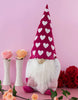 Load image into Gallery viewer, Lifestyle picture of the heart-themed gnome. The gnome stands on a pink table, and the wall behind it is also pink. Two flower vases are placed on the left side of the gnome. One vase has an orange flower, while the other has a pink one. A bouquet of red flowers is placed on the right side of the gnome. Lastly, a stem of peach-colored flowers rests on the table in front of the gnome.
