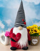 Load image into Gallery viewer, Lifestyle picture of the &quot;Love Bug&quot; gnome. The gnome stands on a wooden table, and the wall behind has a sky wallpaper. On the left, there&#39;s a hollowed circular-shaped flower vase with a pink flower in it. Lastly, on the right, it can be appreciated an oval-shaped basket with a bouquet of sunflowers in it. 
