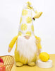 Load image into Gallery viewer, Lifestyle picture of the yellow gnome. It is positioned at a frontal angle, standing on a yellow table. The wall behind is white. In front of the gnome, on the left side of the picture, half of a wired bronze-colored fruit holder with apples in it can be seen. Lastly, next to the gnome on the right side, a white circular base with a decorative lemon on it is visible.
