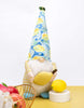 Load image into Gallery viewer, Lifestyle picture of the decorative lemon-themed gnome. It is positioned at a frontal angle, slightly tilted to its left. It is standing on a yellow table with a white wall behind it. In front of the gnome, on the right side, a white circular base with a decorative lemon placed on it is visible. Lastly, on the left side of the picture, half of a wired bronze-colored fruit holder with apples in it can be seen.
