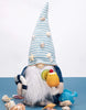 Load image into Gallery viewer, Lifestyle picture of the gnome holding a swim ring. It is positioned from a frontal angle, standing on a blue surface. Many decorative seashells are also placed on the blue surface, surrounding the gnome. Lastly, the background of the picture is white.
