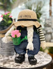Load image into Gallery viewer, Lifestyle photo of the gnome with watering can. It is positioned at a frontal angle, with a slight rightward tilt. The gnome stands on a circular table with a warm gray pattern. Additionally, surrounding the gnome, a garden can be distinguished in a blurred view, with many plants and garden decorations.
