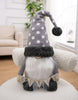 Load image into Gallery viewer, Lifestyle picture of the gray gnome, placed on a white chair covered with faux fur. In the background, white walls, a plant, a window, and beige furniture can be appreciated.
