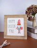 Load image into Gallery viewer, Lifestyle picture of the wooden Easter sign. It is positioned at a frontal angle, slightly tilted to the right, and standing on a brown table in front of a white wall. Next to the plaque, on the right side, there are two stacked books in a diagonal position. On top of the books, there is a cream-colored flower vase with an arrangement of purple flowers. Lastly, two mechanical pencils can be seen placed in its front, one above the other.
