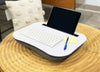 Load image into Gallery viewer, Lifestyle picture of the Rae Dunn lap desk for laptops or paper-based materials. Captured from a slight right angle, the lap desk rests on a circular wooden tabletop. A white keyboard is placed in the center of the desk’s surface. To the right, sticky notes and a pen are visible. Lastly, a tablet is placed in the lap desk’s black slot.
