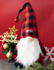 Load image into Gallery viewer, Lifestyle picture of the Christmas plush gnome.  It is placed on a gray surface. There&#39;s a mini Christmas tree behind it, which has a gold-color cutout sign that says &quot;Merry Christmas&quot;.
