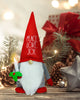 Load image into Gallery viewer, Lifestyle picture of the Rae Dunn Christmas gnome with LED candle.  It is placed on a wooden table, and different Christmas decorations can be appreciated behind the gnome, as pinecones, a gift box. and some Christmas lights.
