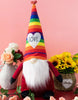 Load image into Gallery viewer, Lifestyle picture of the rainbow theme gnome. Behind it, on the left side, there&#39;s a white flower vase with orange, red, and white flowers. On the right side, also behind the gnome, there&#39;s a basket with sunflowers. The gnome is standing on a red table, and in front of it there are two flower stems. Lastly, the wall behind this arrangement is pink.
