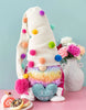 Load image into Gallery viewer, Lifestyle picture of the colorful gnome. Placed at a frontal angle, it stands on a pink table, showcasing a mint-colored wall in the background. On the front left side, a dish with two doughnuts and a piece of Swiss roll can be appreciated. Lastly, on the back right side, there is a white hammered flower vase containing multicolored flowers.
