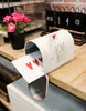 Load image into Gallery viewer, Lifestyle picture of the decorative mailbox with hearts. It is placed at a 45-degree angle on the edge of a wooden table, with a brick wall far behind. The mailbox door is open, revealing a white envelope with illustrated hearts. On the left side, a black plastic plant pot with pink flowers in it can be appreciated. Behind, there&#39;s a black metal desk calendar that marks the date &quot;Mon 14 Feb.&quot; Lastly, below the table, a wooden cabinet can be seen.
