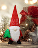 Load image into Gallery viewer, Lifestyle picture of the Santa-theme gnome. It is placed on a wooden table, and different Christmas decorations can be appreciated behind the gnome, as pinecones, a gift box, and some Christmas lights.
