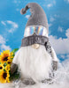 Load image into Gallery viewer, Lifestyle picture of the snowboarder gnome.  It is placed on a surface covered with a white plush tablecloth.  Behind the gnome there&#39;s a wall with a sky-theme  wallpaper on it. Next to it, on its right, there is a sunflower arrangement. 
