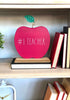 Load image into Gallery viewer, Lifestyle picture of the apple-shaped teacher sign. It is positioned within a shelf at a frontal angle, standing on a book. On the right side of the sign, three books are resting against the side of the shelf. Below, there is another shelf where a white square-shaped desk clock is placed on the left, and four books are resting against the right side.
