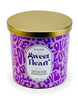 Load image into Gallery viewer, Front angle of the Valentine-themed scented candle. In this view, all the features of the candle can be fully appreciated: The lilac container adorned with a purple-colored pattern of the word &quot;Love,&quot; written with a doodle font, the white label displaying the term &quot;Sweet Heart&quot; and its ingredients; and the golden lid atop the jar. The background of the picture is white.

