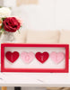 Load image into Gallery viewer, Lifestyle picture of the Valentine-themed sign, positioned in a frontal angle. The sign is placed on a white marble tabletop. Behind the plaque, a flower vase with red and white flowers can be appreciated. Far in the background of the picture, a cream-colored piece of furniture is placed against a white wall.
