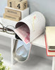 Load image into Gallery viewer, Lifestyle picture of a decorative mailbox with a Valentine theme. It is placed at a 45-degree angle on the edge of a white table. The mailbox door is open, revealing a pink-colored envelope with illustrations of hearts on it. On the left, a pair of glasses can be seen, while two books, one above the other, decorate the right side of the table. The wall behind is gray, and below the table, there&#39;s an empty space.
