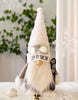 Load image into Gallery viewer, Lifestyle picture of the Rae Dunn winter-themed gnome. It is placed on a surface covered with white plush fabric. It is surrounded by winter and Christmas-themed decorations, as Christmas ornaments and decorative snowflakes. 

