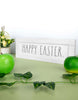 Load image into Gallery viewer, Lifestyle picture of the wooden Easter sign. It is positioned at a frontal angle, slightly tilted to the right, and standing on a sage green table against a white wall. Next to the sign, on the left, there&#39;s a white rounded flower vase with an arrangement of plants and light yellow flowers. In front of the plaque, there are two fruits: a green apple on the left and a green tangerine on the right. Lastly, a leafy stem is positioned in the foreground.
