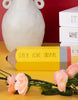 Load image into Gallery viewer, A lifestyle picture of the pencil sign, positioned at a frontal angle on a yellow table. Adorning the scene is a branch with pink flowers resting in front of the pencil. Behind it, a white flower vase can be distinguished. Next to the vase, it can be appreciated a set of three stacked decorative white books. Lastly, the wall far behind is red color.
