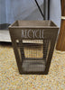 Load image into Gallery viewer, Lifestyle picture of the wooden waste basket, placed at a frontal angle on a gravel floor. In the far background, a gray wall can be seen.
