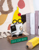 Load image into Gallery viewer, Lifestyle picture of the teacher gnome. It is placed in the center of the picture, standing on a wooden table. It is tilted to the left. Behind the gnome, on the right side, a set of three books can be seen standing on the table. In front of the gnome, also on the right side, a pencil sign with the phrase &quot;Teach, Love, Inspire&quot; rests on the table. A white curtain with red and gold spots is visible in the background.
