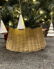 Load image into Gallery viewer, Lifestyle image of the rattan Christmas tree collar. The item is placed on a gray, textured surface, concealing the Christmas tree base. The bottom part of the tree’s branches can be seen, decorated with yellow Christmas bulbs and tree-shaped ornaments. Lastly, various Christmas gift boxes are visible behind the collar.
